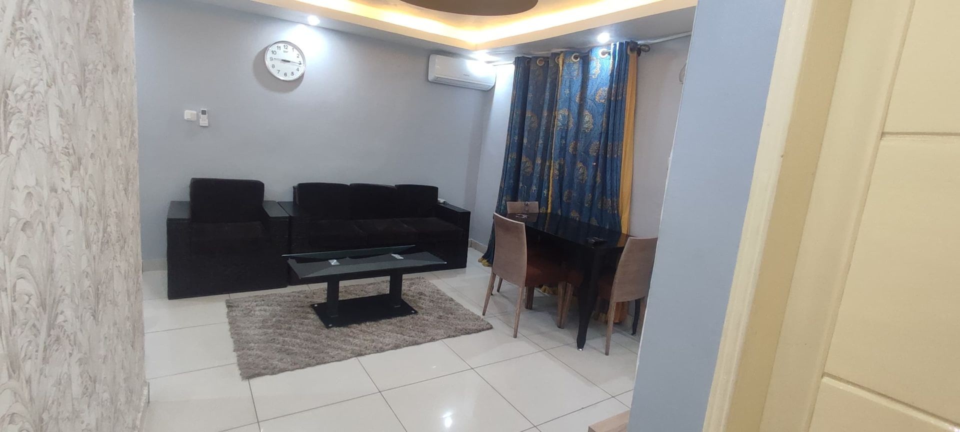 Living room with black sofa, coffee table, dining set, blue curtains, and wall clock.